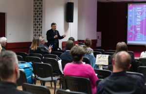 Mężczyzna stoi i mówi do mikrofonu. Uczestnicy konferencji słuchają.