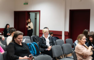 Uczestnicy konferencji siedzą i słuchają. W tle widać stojącą kobietę.