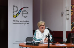 Kobieta wygłasza referat. W tle roll-up Polskiego Seminarium Etnomuzykologicznego.