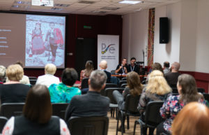 Kobieta i mężczyzna wygłaszają referat. Uczestnicy konferencji siedzą i słuchają. W tle na ekranie prezentacja multimedialna.