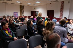 Uczestnicy konferencji siedzą. Kobieta mówi do mikrofonu.